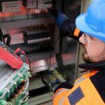 Electrician wearing safety gear operates switchboard in industrial setting.